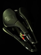 A collection of overlapping black bicycle saddles with textured surfaces and embossed logos. The image is taken against a dark background, highlighting the sleek design and contours of the saddles.