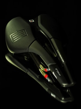A collection of overlapping black bicycle saddles with textured surfaces and embossed logos. The image is taken against a dark background, highlighting the sleek design and contours of the saddles.