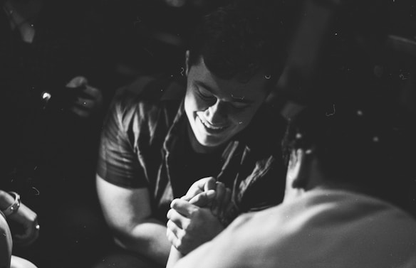 A black and white photograph displaying two people engaging in an arm-wrestling match, with both appearing focused and smiling. The background is blurred, emphasizing the individuals.
