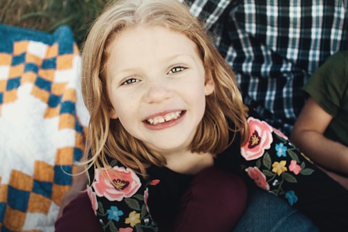 A young girl with blonde hair and a gap-toothed smile is looking up at the camera. She is wearing a black shirt with colorful floral patterns. There's a checkered quilt with orange and blue squares to her left. In the background, a person in a plaid shirt and another in a green shirt are partially visible.