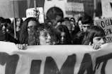 A large group of people, mostly young, are gathered in a protest or demonstration. The crowd holds various signs and banners with text, suggesting a social or environmental cause. Expressions of determination and focus are visible.