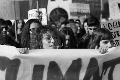 A large group of people, mostly young, are gathered in a protest or demonstration. The crowd holds various signs and banners with text, suggesting a social or environmental cause. Expressions of determination and focus are visible.