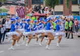 A group of dancers in blue and white costumes perform energetically on a street, surrounded by a parade atmosphere. Spectators can be seen in the background, some wearing vibrant outfits and costumes. A camera crane extends over the dancers, indicating the event is being recorded or broadcasted.