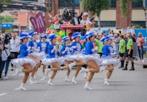 A group of dancers in blue and white costumes perform energetically on a street, surrounded by a parade atmosphere. Spectators can be seen in the background, some wearing vibrant outfits and costumes. A camera crane extends over the dancers, indicating the event is being recorded or broadcasted.