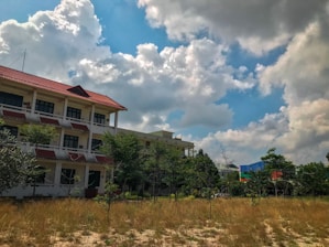A multi-story building with a red tiled roof surrounded by young trees and grass under a partly cloudy sky. The structure features several windows and balconies, and the outdoor area is characterized by sparse vegetation and open land.