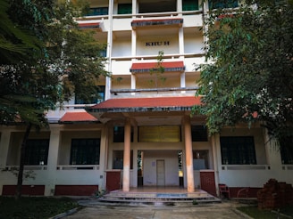 A multi-story building with a red-tiled roof, pillars at the entrance, and surrounded by lush green trees. The structure has several windows, and a sign with 'KHU H' is visible on the upper part of the building. The area in front of the building is a paved path with some grass.