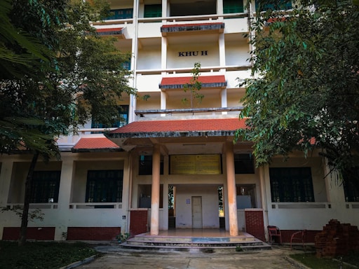 A multi-story building with a red-tiled roof, pillars at the entrance, and surrounded by lush green trees. The structure has several windows, and a sign with 'KHU H' is visible on the upper part of the building. The area in front of the building is a paved path with some grass.