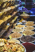 A vibrant market display featuring a variety of dried fruits, nuts, and spices. On the left, shelving units are filled with different kinds of nuts and seeds. In the foreground and to the right, numerous large baskets and bowls showcase an array of colorful dried products, labeled with small yellow price tags. The scene is lively and bustling, with a rich assortment of textures and colors.