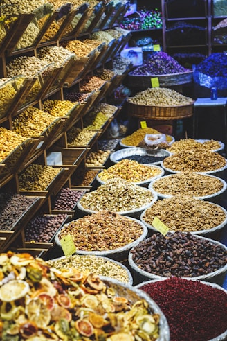A vibrant market display featuring a variety of dried fruits, nuts, and spices. On the left, shelving units are filled with different kinds of nuts and seeds. In the foreground and to the right, numerous large baskets and bowls showcase an array of colorful dried products, labeled with small yellow price tags. The scene is lively and bustling, with a rich assortment of textures and colors.