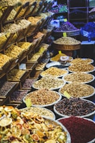 A vibrant market display featuring a variety of dried fruits, nuts, and spices. On the left, shelving units are filled with different kinds of nuts and seeds. In the foreground and to the right, numerous large baskets and bowls showcase an array of colorful dried products, labeled with small yellow price tags. The scene is lively and bustling, with a rich assortment of textures and colors.