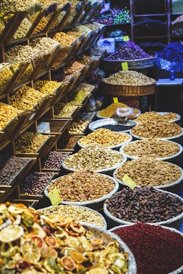 A vibrant market display featuring a variety of dried fruits, nuts, and spices. On the left, shelving units are filled with different kinds of nuts and seeds. In the foreground and to the right, numerous large baskets and bowls showcase an array of colorful dried products, labeled with small yellow price tags. The scene is lively and bustling, with a rich assortment of textures and colors.