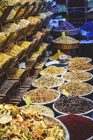 A vibrant market display featuring a variety of dried fruits, nuts, and spices. On the left, shelving units are filled with different kinds of nuts and seeds. In the foreground and to the right, numerous large baskets and bowls showcase an array of colorful dried products, labeled with small yellow price tags. The scene is lively and bustling, with a rich assortment of textures and colors.