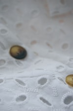 A close-up view of a fabric with a delicate embroidery pattern featuring oval cutouts and stitching on a soft, white background. Two buttons with a sheen, one black and one gold, are sewn onto the fabric, adding subtle contrast.