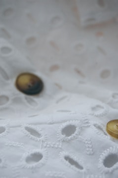 A close-up view of a fabric with a delicate embroidery pattern featuring oval cutouts and stitching on a soft, white background. Two buttons with a sheen, one black and one gold, are sewn onto the fabric, adding subtle contrast.