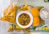 A traditional Indian meal is presented on a table, featuring a bowl of spicy curry garnished with herbs. Accompanying the dish is a plate with two buns, a bowl of chopped onions, and a bag of crispy snacks. A spoon is placed nearby on the tablecloth.