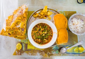 A traditional Indian meal is presented on a table, featuring a bowl of spicy curry garnished with herbs. Accompanying the dish is a plate with two buns, a bowl of chopped onions, and a bag of crispy snacks. A spoon is placed nearby on the tablecloth.