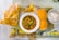 A traditional Indian meal is presented on a table, featuring a bowl of spicy curry garnished with herbs. Accompanying the dish is a plate with two buns, a bowl of chopped onions, and a bag of crispy snacks. A spoon is placed nearby on the tablecloth.