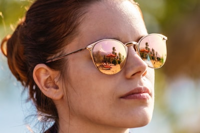 A person wearing reflective sunglasses that show a beach scene with palm trees and a structure in the reflection. The background is slightly blurred, emphasizing the focus on the reflective surface of the sunglasses.