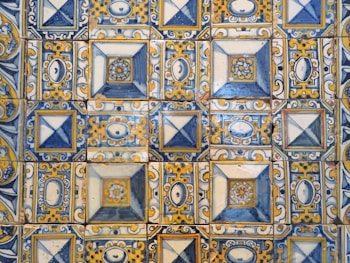 A detailed arrangement of ornately patterned ceramic tiles featuring geometric designs and floral motifs in a combination of blue, yellow, and white hues. Each tile is bordered with intricate swirling patterns, and some tiles have diamond or oval shapes centered within squares.