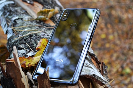 A smartphone rests against a broken tree branch, surrounded by autumn leaves. The phone's shiny surface reflects the blurred background, which includes shades of brown, yellow, and green.