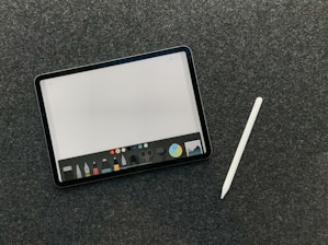 A digital tablet with a stylus rests on a dark textured surface. The tablet screen displays a drawing or writing application interface, featuring a variety of tool icons and color options at the bottom.