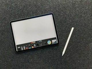 A digital tablet with a stylus rests on a dark textured surface. The tablet screen displays a drawing or writing application interface, featuring a variety of tool icons and color options at the bottom.