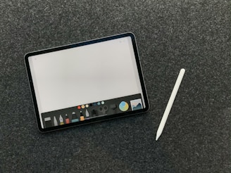 A digital tablet with a stylus rests on a dark textured surface. The tablet screen displays a drawing or writing application interface, featuring a variety of tool icons and color options at the bottom.