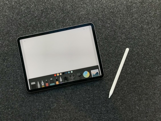 A digital tablet with a stylus rests on a dark textured surface. The tablet screen displays a drawing or writing application interface, featuring a variety of tool icons and color options at the bottom.