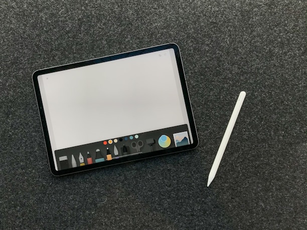 A digital tablet with a stylus rests on a dark textured surface. The tablet screen displays a drawing or writing application interface, featuring a variety of tool icons and color options at the bottom.