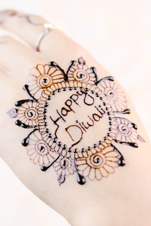 A hand adorned with intricate henna designs featuring floral and circular patterns. The centerpiece of the design includes the words 'Happy Diwali' written in an artistic style. The artwork is detailed, with delicate spirals and dots enhancing the visual appeal, and a ring is visible on one of the fingers.