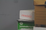 A stack of cardboard boxes, with the top box labeled 'FRAGILE' in red text. Below it, a green box marked 'Sustainable Earth' is visible. Another box, brown with black stripes, is partially in view on the right.