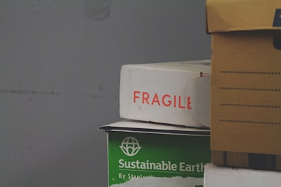 A stack of cardboard boxes, with the top box labeled 'FRAGILE' in red text. Below it, a green box marked 'Sustainable Earth' is visible. Another box, brown with black stripes, is partially in view on the right.