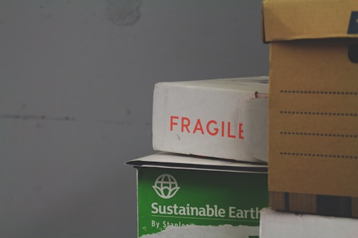 A stack of cardboard boxes, with the top box labeled 'FRAGILE' in red text. Below it, a green box marked 'Sustainable Earth' is visible. Another box, brown with black stripes, is partially in view on the right.