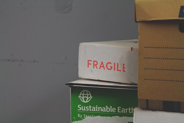 A stack of cardboard boxes, with the top box labeled 'FRAGILE' in red text. Below it, a green box marked 'Sustainable Earth' is visible. Another box, brown with black stripes, is partially in view on the right.