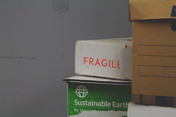 A stack of cardboard boxes, with the top box labeled 'FRAGILE' in red text. Below it, a green box marked 'Sustainable Earth' is visible. Another box, brown with black stripes, is partially in view on the right.