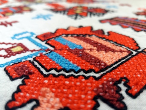 A detailed close-up of a colorful embroidered pattern featuring geometric shapes. The design includes vibrant reds, blues, and soft pinks, outlined with black stitching on a light fabric background. The texture of the embroidery is prominently visible, showing fine details of the individual threads.