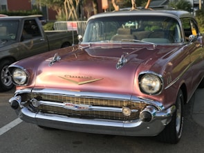 A classic Chevrolet car with a vintage design, featuring a shiny chrome front grille and headlight bezels. The car has a sleek, metallic pink exterior and sits parked on a street with another vehicle partially visible in the background.