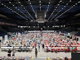 An indoor sports arena filled with rows of tables, each occupied by people engaged in competitive activities. The ceiling is high, with numerous lights illuminating the large space. The audience seating on either side is empty, and there are flags hanging from the upper level.