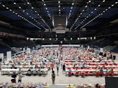 An indoor sports arena filled with rows of tables, each occupied by people engaged in competitive activities. The ceiling is high, with numerous lights illuminating the large space. The audience seating on either side is empty, and there are flags hanging from the upper level.