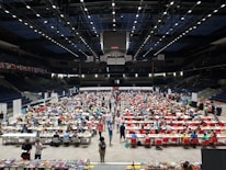 An indoor sports arena filled with rows of tables, each occupied by people engaged in competitive activities. The ceiling is high, with numerous lights illuminating the large space. The audience seating on either side is empty, and there are flags hanging from the upper level.