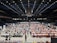 An indoor sports arena filled with rows of tables, each occupied by people engaged in competitive activities. The ceiling is high, with numerous lights illuminating the large space. The audience seating on either side is empty, and there are flags hanging from the upper level.