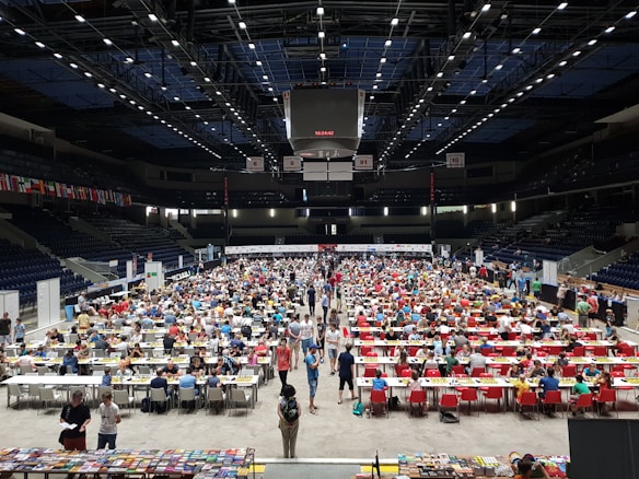 An indoor sports arena filled with rows of tables, each occupied by people engaged in competitive activities. The ceiling is high, with numerous lights illuminating the large space. The audience seating on either side is empty, and there are flags hanging from the upper level.