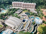 An aerial view of a large resort or theme park featuring multiple pools, water slides, and various structures surrounded by lush greenery. Central to the image is a large building with a distinctive roof, and beside it, several smaller buildings and open spaces, including a turquoise pool area and colorful water slides.