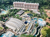 An aerial view of a large resort or theme park featuring multiple pools, water slides, and various structures surrounded by lush greenery. Central to the image is a large building with a distinctive roof, and beside it, several smaller buildings and open spaces, including a turquoise pool area and colorful water slides.