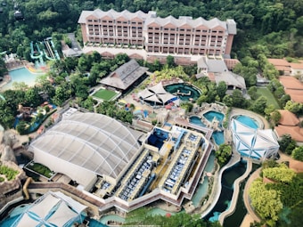 An aerial view of a large resort or theme park featuring multiple pools, water slides, and various structures surrounded by lush greenery. Central to the image is a large building with a distinctive roof, and beside it, several smaller buildings and open spaces, including a turquoise pool area and colorful water slides.