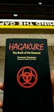 A person holds a book titled 'Hagakure: The Book of the Samurai' by Yamamoto Tsunetomo, translated by William Scott Wilson. The book cover features a red symbol on a black background. Behind the book, a subway platform edge is visible with the words 'Mind the Gap' painted on it.