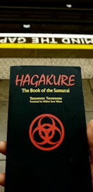 A person holds a book titled 'Hagakure: The Book of the Samurai' by Yamamoto Tsunetomo, translated by William Scott Wilson. The book cover features a red symbol on a black background. Behind the book, a subway platform edge is visible with the words 'Mind the Gap' painted on it.