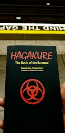 A person holds a book titled 'Hagakure: The Book of the Samurai' by Yamamoto Tsunetomo, translated by William Scott Wilson. The book cover features a red symbol on a black background. Behind the book, a subway platform edge is visible with the words 'Mind the Gap' painted on it.