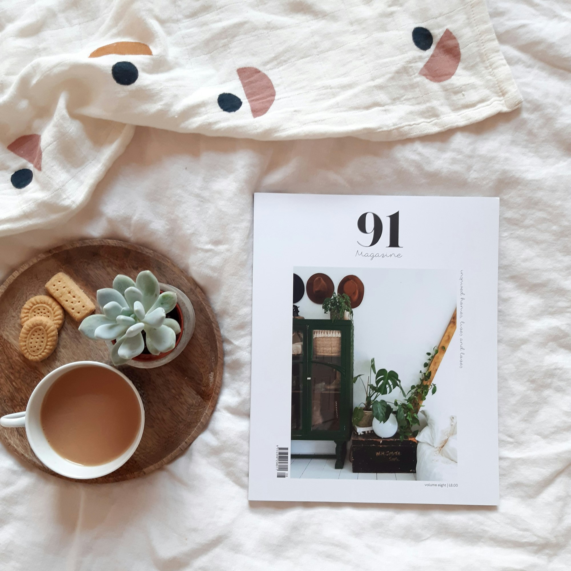 a print magazine about interiors and lifestyle on a bed with a cup of tea and biscuits and succulent on a tray. 