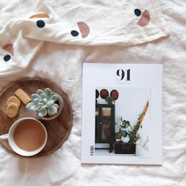 A white magazine cover with the title '91 Magazine' on a soft white blanket, alongside a wooden tray containing a cup of coffee, cookies, and a small succulent plant.